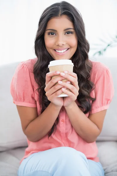 Cheerful cute brunette sitting on couch holding disposable cup — Stock Photo, Image