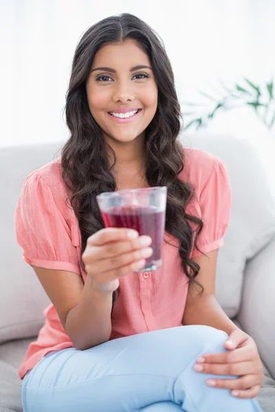 Cheerful cute brunette sitting on couch holding glass of juice — Stock Photo, Image