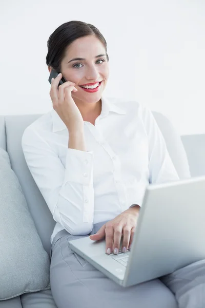 Mujer bien vestida usando portátil y teléfono celular en el sofá — Foto de Stock