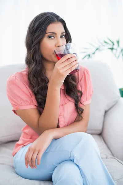 Zufriedene süße Brünette sitzt auf der Couch und trinkt ein Glas Saft — Stockfoto