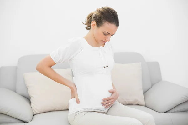 Worried pregnant woman touching her injured back sitting on couch — Stock Photo, Image