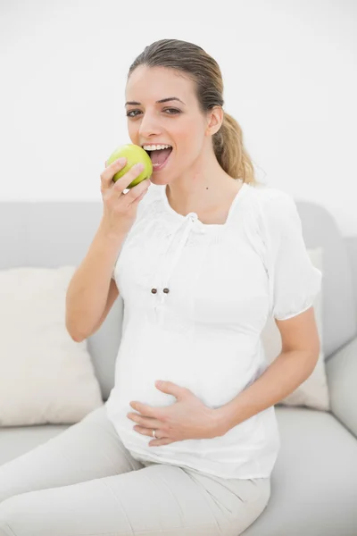 Content pregnant woman eating green apple sitting on couch — Stock Photo, Image
