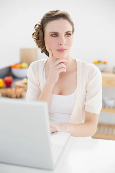 Mujer linda pensativa sentada en la cocina usando su computadora portátil —  Fotos de Stock