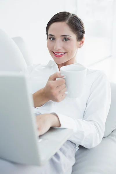 Donna sorridente ben vestita con laptop e tazza di caffè sul divano — Foto Stock