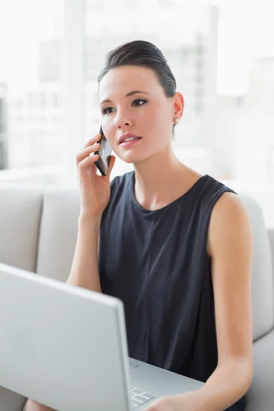 Beautiful well dressed woman using laptop and cellphone on sofa — Stock Photo, Image