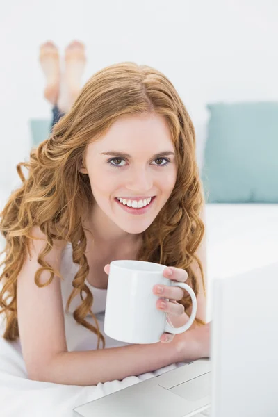 Kızıl saçlı kız yatakta dizüstü kullanarak kahve fincanı ile gülümseyen — Stok fotoğraf