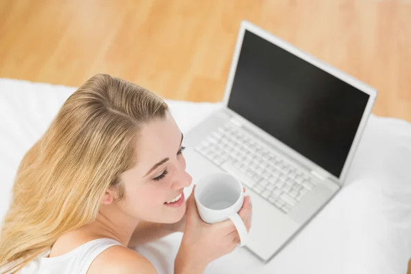 Mujer rubia calma sosteniendo una taza acostada en la cama junto a su computadora portátil —  Fotos de Stock
