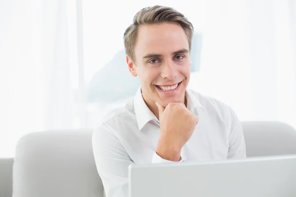 Usmívající se neformální mladý muž pomocí přenosného počítače na pohovce — Stock fotografie