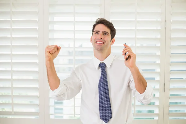 Cheerful businessman with mobile phone clenching fist in office — Stock Photo, Image