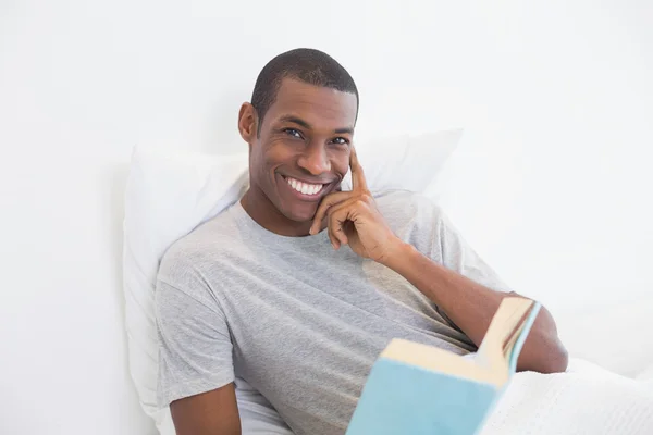 Jeune homme afro souriant avec un livre au lit — Photo