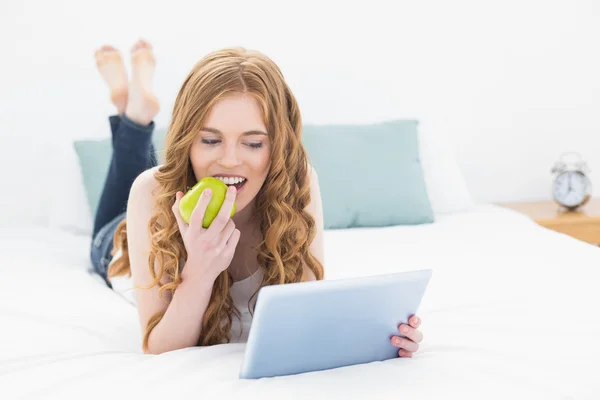 按住一个苹果并在床上使用 tablet pc 的休闲红头发的女孩 — 图库照片