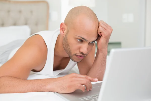 Serious casual bald young man using laptop in bed — Stock Photo, Image