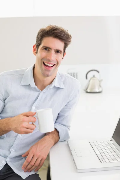 Sonriente hombre casual con taza de café con portátil en casa — Foto de Stock