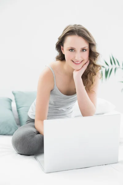 Sonriente morena casual con portátil sentado en la cama — Foto de Stock