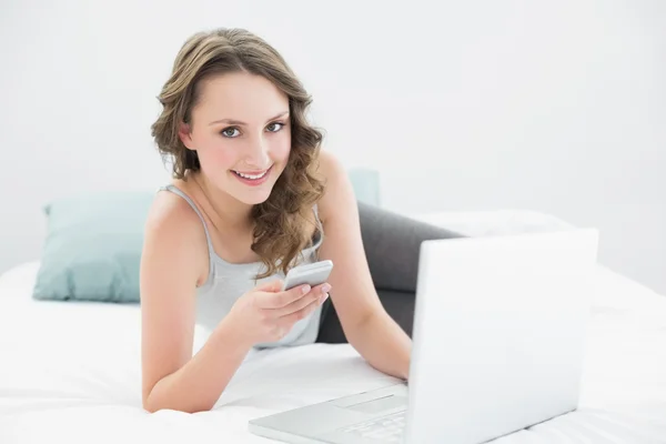 Smiling casual woman with cellphone and laptop in bed — Stock Photo, Image