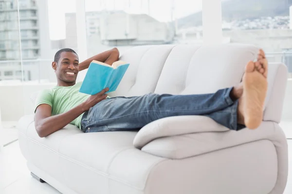 Ritratto a figura intera di un uomo afro sorridente che legge un libro sul divano — Foto Stock