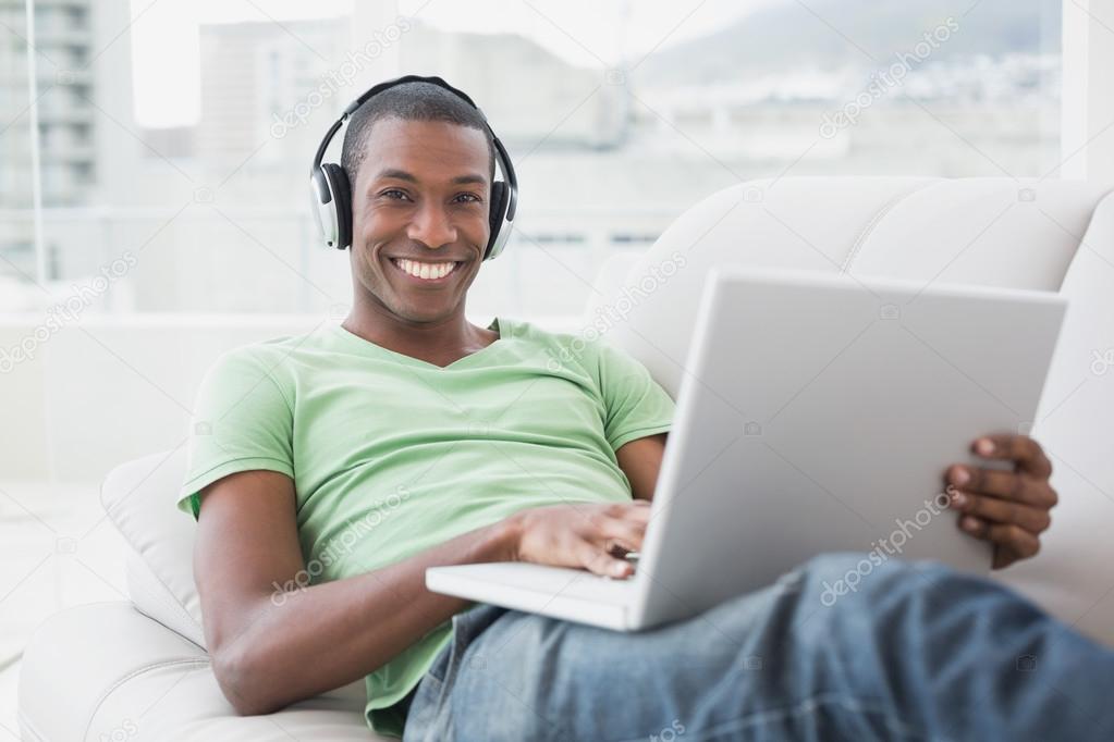 Relaxed smiling Afro man with headphones using laptop on sofa