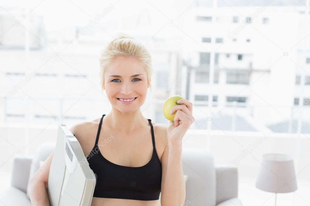 Smiling fit woman holding scale and apple in fitness studio