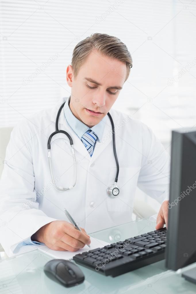 Doctor writing a note while using computer at medical office