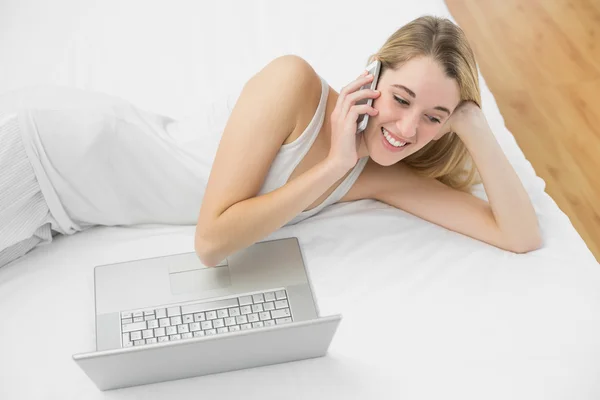 Mujer rubia encantadora llamando con su teléfono inteligente tumbado en su cama — Foto de Stock