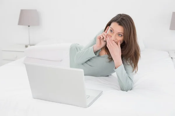 Mujer casual conmocionada usando el teléfono celular y el portátil en la cama —  Fotos de Stock