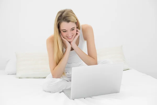 Vrolijk jonge vrouw die werkt met haar laptop zittend op haar bed — Stockfoto