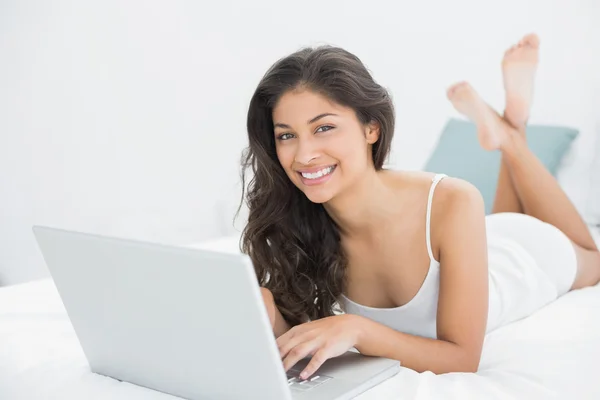 Sonriente mujer joven casual usando el ordenador portátil en la cama —  Fotos de Stock