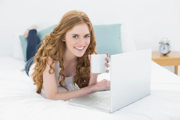 Sonriente chica pelirroja con taza de café usando el ordenador portátil en la cama —  Fotos de Stock