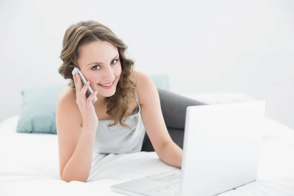 Leende casual kvinna med mobiltelefon och bärbar dator i sängen — Stockfoto