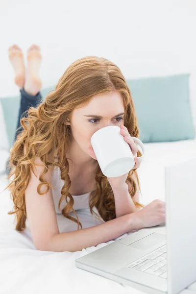 休闲的金发女郎在床上使用笔记本电脑时喝咖啡 — 图库照片