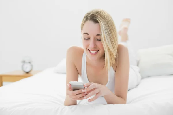 Mujer joven contenta enviando mensajes de texto con su teléfono inteligente acostado en su cama —  Fotos de Stock