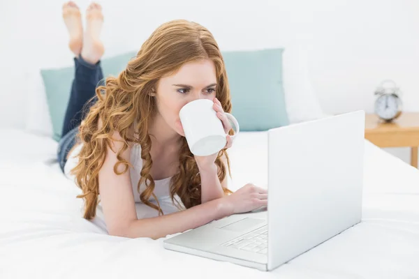 Biondo casuale bere caffè mentre si utilizza il computer portatile a letto — Foto Stock