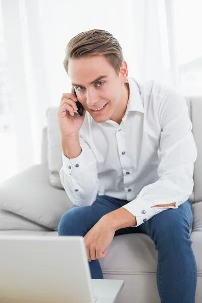 Hombre joven casual utilizando el teléfono celular y el ordenador portátil en el sofá — Foto de Stock