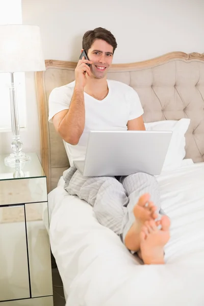 Casual χαμογελαστός άνθρωπος χρησιμοποιώντας το κινητό και το laptop στο κρεβάτι — Φωτογραφία Αρχείου