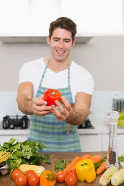 Smiling man holding out bell pepper with vegetables in kitchen — Stock Photo, Image