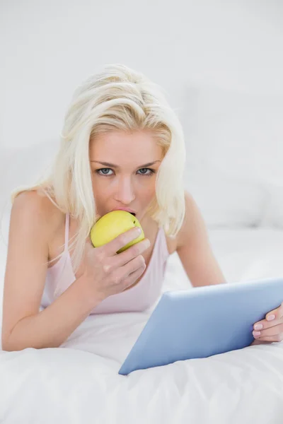 Lässige Blondine isst einen Apfel, während sie den Tablet-PC im Bett benutzt — Stockfoto