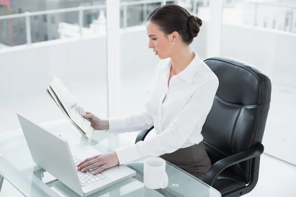 Businesswoman reading newspaper and using laptop in office — Stock Photo, Image