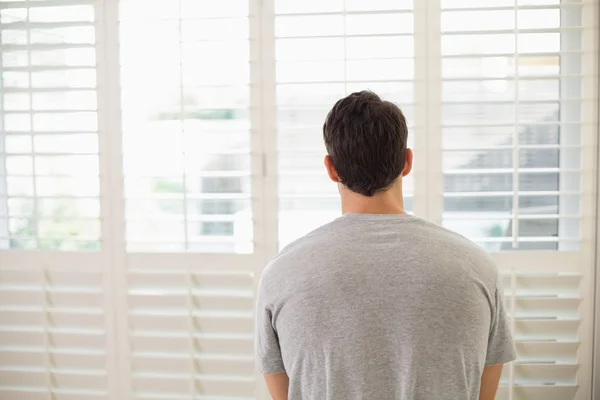 Vista trasera del hombre mirando a través de persianas de la ventana en la habitación luminosa —  Fotos de Stock
