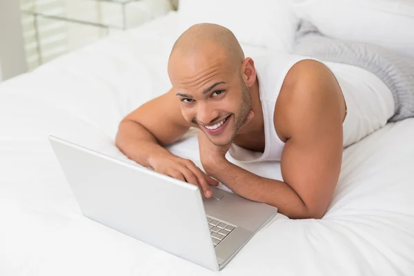 Usmívající se příležitostné holohlavý mladík pomocí notebooku v posteli — Stock fotografie