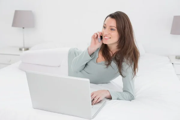 Sorridente donna casual utilizzando il cellulare e il computer portatile a letto — Foto Stock