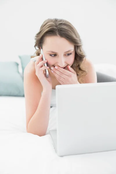 Smiling casual woman using cellphone and laptop in bed — Stock Photo, Image