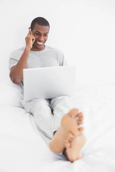 Afro man using cellphone and laptop in bed — Stock Photo, Image