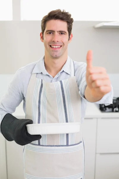 Человек с блюдом для выпечки жестикулирует большие пальцы вверх на кухне — стоковое фото