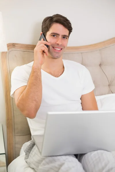 Hombre sonriente casual usando teléfono celular y portátil en la cama —  Fotos de Stock