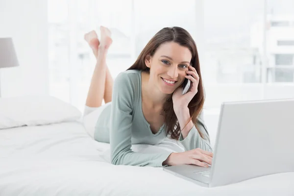 Smiling casual woman using cellphone and laptop in bed — Stock Photo, Image