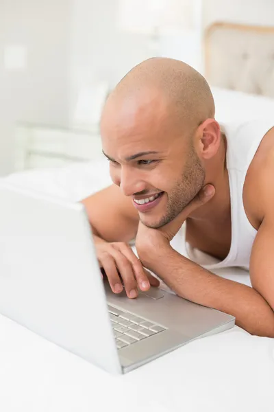 Smiling casual bald young man using laptop in bed