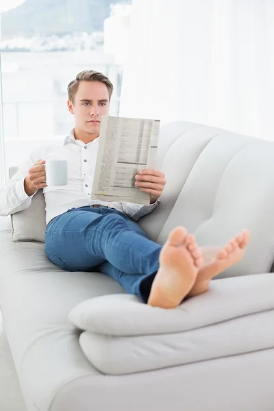 Relajado hombre bebiendo café mientras lee el periódico en el sofá — Foto de Stock