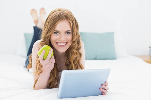 Casual red-haired girl using tablet PC while holding an apple in bed — Stock Photo, Image