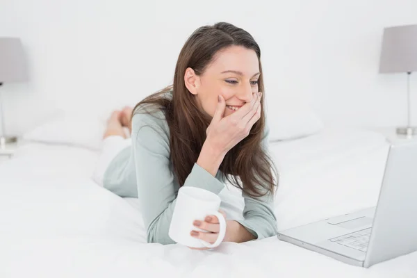Fröhliche Frau mit Laptop und Tasse im Bett — Stockfoto