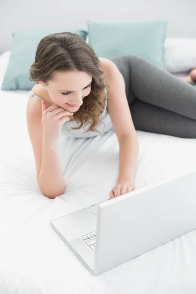 Smiling casual young brunette with laptop in bed — Stock Photo, Image